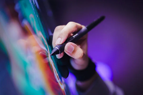 Close-up of a hand holding a digital stylus, drawing vibrant artwork on a tablet with a colorful and blurred background.tutorials